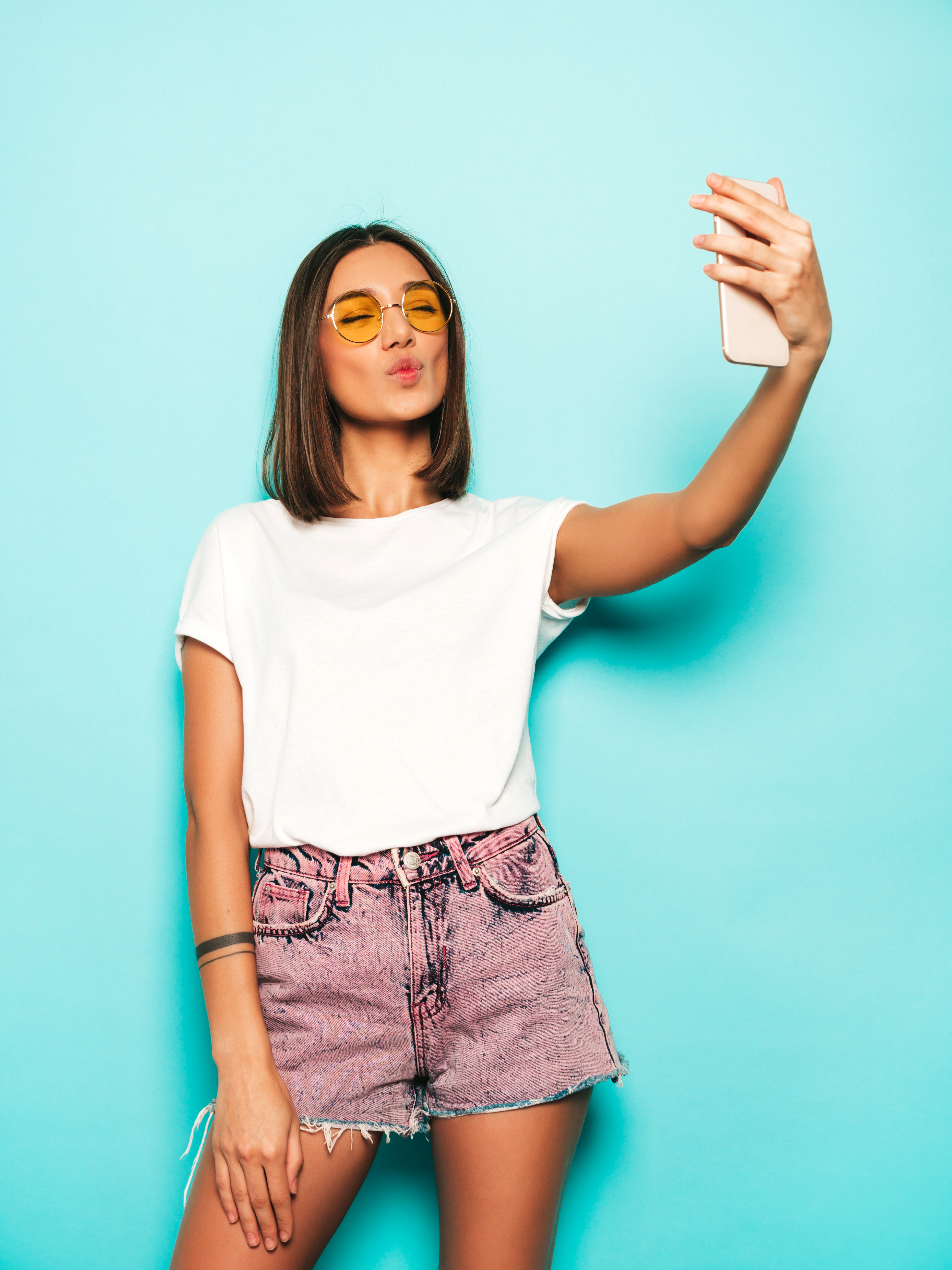 hermosa-modelo-sonriente-vestida-ropa-hipster-verano-chica-despreocupada-sexy-posando-estudio-junto-pared-azul-shorts-jeans-mujer-moda-divertida-tomando-fotos-autorretrato-selfie-telefono-inteligente